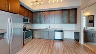 This single-floor residence showcases a thoughtfully designed open-concept layout with consistent hardwood-style flooring flowing throughout. The living area is a standout feature, highlighted by expansive floor-to-ceiling windows that frame sweeping city skyline views, flooding the space with abundant natural light and creating a strong visual connection to the urban landscape beyond.

The kitchen is well-appointed with rich, dark wood upper cabinetry featuring glass-front panels, complemented by lower gray cabinetry. A full suite of stainless steel appliances includes a refrigerator with ice maker, range, microwave, and dishwasher. Track lighting overhead illuminates the workspace, while light-toned countertops provide ample preparation surface.

A hallway connects the living areas to additional rooms, offering a closet with wire shelving for organized storage. The bathroom is finished with a granite-topped vanity set in dark cabinetry, a large framed mirror with vanity lighting, a soaking tub and shower combination with decorative tile accents, and a wall-mounted cabinet for additional storage.

Overall, the unit presents a clean, contemporary aesthetic with a neutral color palette, combining functional design with elevated finishes throughout.