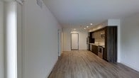 This single-floor space showcases a clean, contemporary design with an open-concept layout that seamlessly connects the living area to the kitchen. Light-toned hardwood-style flooring runs throughout, lending warmth and visual continuity across the space. The high, white walls contribute to an airy, expansive feel, while recessed ceiling lights provide bright, even illumination throughout.

The kitchen is positioned along one wall and is equipped with stainless steel appliances, including a refrigerator, range, and microwave. Rich, dark cabinetry offers a striking contrast against the light walls and flooring, and a sink with countertop workspace completes the functional layout. The kitchen's design reflects a modern aesthetic with efficient use of space.

A doorway visible from the main living area leads to additional rooms or utility spaces, suggesting a practical and well-organized floor plan. A hallway along one side of the unit hints at further private areas beyond the open living zone. Ceiling vents indicate a central HVAC system. Overall, the space exudes a minimalist, move-in-ready ambiance suited to contemporary living.