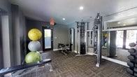 This fitness center offers a compact yet fully functional workout environment designed to support a variety of exercise routines. The space is finished with durable, dark-toned carpet tiles that provide cushioning underfoot and a practical surface suited for high-traffic athletic use. The walls are painted in a modern charcoal gray, giving the room a focused and energetic atmosphere enhanced by recessed ceiling lighting that ensures consistent illumination throughout.

The strength training area is anchored by a multi-functional cable machine, which supports a wide range of resistance exercises for both upper and lower body conditioning. Adjustable weight benches are positioned nearby, offering flexibility for various lifting and toning routines. A collection of stability and exercise balls in multiple sizes is neatly stacked in the corner, providing options for balance training, core work, and stretching. Wall-mounted mirrors along one side of the room expand the sense of space and allow users to monitor their form during workouts.

An exterior door with a window allows for natural light to filter in, adding a sense of openness to the room. The single-floor layout ensures straightforward accessibility, making this fitness center a practical and well-appointed facility for everyday exercise needs.