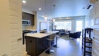 This contemporary single-floor space showcases a thoughtfully designed open-concept layout that effortlessly connects the kitchen and living areas. The kitchen is a standout feature, boasting dark espresso cabinetry, stainless steel appliances including a gas range and over-the-range microwave, and a striking central island topped with a crisp white countertop. Two modern pendant lights hang above the island, adding both style and functional task lighting. Bar-height seating at the island creates a casual dining and gathering spot.

The kitchen flows naturally into the living area, which is anchored by a large picture window that invites an abundance of natural light and offers views of the surrounding greenery. The light hardwood flooring throughout unifies the space and contributes to its warm, modern aesthetic. Recessed ceiling lighting complements the natural light, ensuring the entire area remains bright and welcoming at any time of day.

The living area is generously proportioned, providing ample room for comfortable furnishings and entertaining. Clean lines, a neutral color palette, and high-quality finishes throughout give this space a polished, contemporary feel that balances both style and everyday functionality.