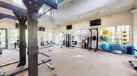 Located at 1021 Glenn Valley Lane in Indian Trail, NC, this fitness center offers a thoughtfully designed exercise room that provides an inviting and functional environment for a comprehensive workout experience. The space features soaring two-story ceilings with a dramatic coffered skylight detail, flooding the interior with natural light alongside an array of large windows positioned on multiple walls. The floors are finished with patterned carpet tiles that add a clean, modern aesthetic throughout.

The exercise room is comprehensively equipped for both cardio and strength training. Cardio options include treadmills, stair climbers, stationary bikes, and rowing machines, many featuring personal display screens. Strength training is well-supported with a multi-station cable machine, a dedicated dumbbell rack with a full range of free weights, a weight bench, and a pull-up and dip tower station. A functional training corner includes stability balls, medicine balls, resistance bands, and balance equipment, supporting flexibility and rehabilitation exercises.

Large wall-mounted mirrors enhance the sense of space and allow users to monitor their form, while multiple wall-mounted flat-screen televisions provide entertainment throughout the room. Recessed lighting and crown molding add a polished, upscale finish to the space, making this fitness center a well-rounded facility suited for a wide variety of training goals.