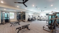 This fitness center is designed to support a full spectrum of workout routines within a spacious, single-floor layout. The space is finished with neutral-toned carpet flooring and light gray walls, creating a clean and modern aesthetic throughout. Recessed lighting is complemented by multiple ceiling fans, ensuring the space remains well-lit and comfortable during use.

The cardio zone features a row of Matrix treadmills, elliptical trainers, a rowing machine, and a stationary bike, each equipped with individual display screens for interactive workout tracking. Alongside these, a dedicated strength training area houses Matrix cable and functional training machines, providing versatile resistance options for users of all fitness levels.

A free weights section offers a full rack of dumbbells alongside adjustable benches and a large wall-mounted flat-screen TV. Full-length mirrors line portions of the walls, enhancing the sense of space and allowing users to monitor their form. A functional training zone includes a multi-station rig with a heavy punching bag and battle ropes, ideal for high-intensity interval training.

Natural light filters in through multiple windows, offering views of the surrounding outdoor greenery. A wall-mounted water fountain station provides convenient hydration access. The overall ambiance is professional, bright, and inviting, making this fitness center a comprehensive facility for diverse training needs.