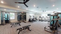 This fitness center is designed to support a full spectrum of workout routines within a spacious, single-floor layout. The space is finished with neutral-toned carpet flooring and light gray walls, creating a clean and modern aesthetic throughout. Recessed lighting is complemented by multiple ceiling fans, ensuring the space remains well-lit and comfortable during use.

The cardio zone features a row of Matrix treadmills, elliptical trainers, a rowing machine, and a stationary bike, each equipped with individual display screens for interactive workout tracking. Alongside these, a dedicated strength training area houses Matrix cable and functional training machines, providing versatile resistance options for users of all fitness levels.

A free weights section offers a full rack of dumbbells alongside adjustable benches and a large wall-mounted flat-screen TV. Full-length mirrors line portions of the walls, enhancing the sense of space and allowing users to monitor their form. A functional training zone includes a multi-station rig with a heavy punching bag and battle ropes, ideal for high-intensity interval training.

Natural light filters in through multiple windows, offering views of the surrounding outdoor greenery. A wall-mounted water fountain station provides convenient hydration access. The overall ambiance is professional, bright, and inviting, making this fitness center a comprehensive facility for diverse training needs.