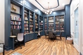 Located at 4285 Johns Creek Pkwy in Suwanee, Georgia, this single-floor space is a beautifully appointed library and office that blends classic sophistication with functional design. The room is defined by dramatic floor-to-ceiling built-in bookshelves finished in a deep navy-blue, flanking all walls and offering extensive storage with granite-topped lower cabinets. The shelves are stocked with books and tasteful decorative accents, creating an immersive, scholarly atmosphere. At the center of the room, a round table with tufted leather chairs serves as a collaborative workspace, while a dedicated desk area with a computer station is seamlessly integrated into the built-ins along one wall. Warm wood-look tile flooring runs throughout, providing an inviting contrast to the bold dark cabinetry. Crown molding and a coffered ceiling detail frame an elegant chandelier that anchors the room with refined lighting. The seating area features a tufted leather sofa, an accent chair, and a patterned area rug beneath textured wallpaper, adding depth and comfort to the space. Natural light filters in through blinds-fitted windows near the entry, complemented by recessed lighting. A glass-paneled door provides a bright and welcoming entry point into this distinguished space.