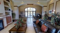 This single-floor space presents a well-appointed lobby or reception area distinguished by its vaulted ceiling with decorative coffered detailing and recessed lighting. The architectural centerpiece is a striking arched entryway fitted with leaded, stained-glass double doors that allow natural light to filter into the interior, creating a bright and inviting atmosphere. Light-toned tile flooring extends throughout the space, enhancing the sense of openness.

The seating area features a dark leather sofa accented with warm-toned decorative pillows, complemented by a solid wood coffee table adorned with a lush floral arrangement. Additional seating is arranged toward the front of the space near the entry doors. Lush tropical plants and decorative botanicals are positioned throughout, adding vibrancy and a welcoming ambiance.

Toward the rear, a reception or concierge desk is staffed and positioned to greet visitors upon entry. Framed artwork on the walls adds a finishing touch. The overall design blends traditional architectural elements with comfortable furnishings, conveying a professional yet approachable environment suitable for a community clubhouse, office, or hospitality setting.