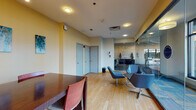 This single-floor professional office space is thoughtfully designed to accommodate both welcoming visitors and conducting business. The layout includes a casual lounge area furnished with contemporary swivel chairs and a round side table, ideal for informal meetings or waiting. Warm yellow accent walls contrast with neutral tones throughout, creating an inviting and professional atmosphere. Light wood-look flooring runs throughout the space, lending warmth and continuity across all areas.

A dedicated meeting or conference area features a substantial dark wood table surrounded by cognac-colored chairs, illuminated by a sleek pendant light fixture. Floor-to-ceiling windows frame lush outdoor views, flooding the room with natural light and providing a pleasant backdrop for meetings.

The space also incorporates glass partition walls that connect to an adjacent lobby area, enhancing openness and visibility between zones. Abstract teal and gold artwork adorns the walls, adding a refined, curated aesthetic. Practical details such as recessed and flush-mount lighting, HVAC vents, and private-access doors ensure the space is both functional and polished.

The overall ambiance balances professionalism with approachability, making this office well-suited for client-facing operations, leasing services, or administrative functions.