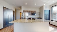 This single-floor unit offers a well-designed open layout with a cohesive blend of modern finishes and functional design. The kitchen serves as a central highlight, anchored by a generously sized island topped with light granite countertops and an undermount sink. Dark-toned cabinetry with sleek hardware lines the walls, accompanied by open shelving, stainless steel appliances including a refrigerator, range, and over-the-range microwave. Light wood-style flooring runs through the main living and kitchen areas, adding warmth to the neutral palette. A large window in the dining area fills the space with natural light and provides views of the surrounding neighborhood. The bedroom is carpeted, offering a softer feel underfoot, and features a ceiling fan and a window that brings in additional daylight. The primary bathroom includes a dual-sink vanity with granite countertops, dark cabinetry, wood-look flooring, a walk-in closet, and a separate shower. A secondary bathroom provides a single-sink vanity with matching granite surfaces and a tub-shower combination. Recessed lighting, dark-stained craftsman-style interior doors, and consistent granite and cabinetry selections throughout contribute to a cohesive, contemporary aesthetic.