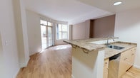 This single-floor residence offers a thoughtfully designed open-concept layout that seamlessly connects the kitchen and living area. The living space features warm hardwood flooring, neutral taupe accent walls, and large floor-to-ceiling sliding glass doors that flood the room with natural light and provide direct access to a private balcony with outdoor views.

The kitchen is positioned behind a raised peninsula counter topped with granite-style stone, providing both workspace and bar seating. Light maple cabinetry lines the kitchen walls, complemented by stainless steel appliances including a range, built-in microwave, and refrigerator. Recessed lighting illuminates the kitchen area, enhancing its clean, modern aesthetic.

A hallway leads to the bedroom, which continues the natural light theme with its own set of sliding glass balcony doors and is finished with plush wall-to-wall carpeting. The hallway also provides access to an in-unit laundry area, adding everyday convenience.

The bathroom is well-appointed with a generous vanity featuring a granite-topped countertop, light maple cabinetry with brushed nickel hardware, an oversized mirror with overhead lighting, and a full tub-shower combination. Hardwood-style flooring ties the space together cohesively throughout the non-carpeted areas, creating a clean and consistent aesthetic across the entire residence.