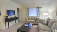 This single-floor residence offers a functional and inviting layout designed for comfortable everyday living. The living room serves as the heart of the home, featuring soft, neutral-toned carpeting that adds warmth underfoot and a light cream color scheme on the walls that creates a bright, airy atmosphere. A plush beige sofa and loveseat are arranged around a dark espresso coffee table, providing a cozy central gathering space. Natural light filters in through a window dressed with decorative blue patterned curtains and horizontal blinds, lending the room a pleasant, well-lit ambiance. A wall-mounted flat-screen television is positioned across from the seating area atop a sleek dark console table, making the space ideal for both relaxing and entertaining. A floor lamp in the corner adds supplemental warm lighting. An interior door visible from the living area provides access to additional rooms, hinting at a thoughtfully connected floor plan. Overall, the space combines practical design with a clean, understated aesthetic, making it a welcoming and versatile environment.