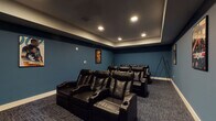 This dedicated home theater is designed to deliver an immersive cinematic experience. The room is finished with deep teal walls accented by a dark coffered ceiling border, creating a dramatic, theater-like ambiance. Recessed lighting is strategically placed to provide subtle illumination without interfering with screen visibility. The flooring is covered in a patterned, dark-toned carpet that complements the room's overall color palette and helps with acoustic dampening.

Seating is arranged in two tiered rows of black leather power recliners, each equipped with built-in cup holders, ensuring comfort during extended viewing sessions. The arrangement provides clear sightlines to the large wall-mounted flat-screen display positioned at the front of the room. Flanking the screen, a decorative carved wood sideboard offers additional storage and display space, while a tripod-style speaker stand contributes to the room's audio setup.

Classic movie posters—including nods to Top Gun, Indiana Jones, and James Bond—are framed and displayed along the walls, reinforcing the cinematic theme. A single entry door with a paneled design provides access to the room, and subtle in-ceiling speakers enhance the audio experience. Overall, the space is thoughtfully designed for dedicated entertainment use.