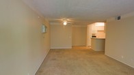 This single-floor residence offers a straightforward and functional layout designed for comfortable everyday living. The main living area is generously sized, featuring wall-to-wall carpeting in a neutral beige tone that adds warmth underfoot. A ceiling fan with a light fixture is centrally mounted on the textured ceiling, providing both ambient lighting and air circulation throughout the space. The walls are finished in a soft, neutral tone that complements the overall understated aesthetic.

Seamlessly connected to the living area is the kitchen, which is open to the main space and separated by a raised breakfast bar counter — ideal for casual dining or entertaining. The kitchen is equipped with white cabinetry, a microwave, and additional appliances, all set beneath warm overhead lighting. The white finish of the cabinetry brightens the kitchen area and contrasts neatly with the darker countertop surfaces.

An electrical panel is conveniently accessible on one of the living room walls, and the layout flows efficiently throughout the single level. The overall ambiance of the space is clean, neutral, and move-in ready, making it a practical and welcoming environment suited to a variety of living arrangements.