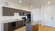 This thoughtfully designed one-bedroom, one-bathroom unit delivers a functional and modern living experience across a single floor. The kitchen is a standout feature, showcasing dark espresso cabinetry, light countertops, and a full suite of appliances including a stainless steel refrigerator, black electric range, built-in microwave, and dishwasher. A central island with a dark granite-style surface provides additional prep space and casual seating. Light wood-style flooring extends throughout the main living areas, creating a warm and cohesive aesthetic. The open layout connects the kitchen seamlessly to the living room, which benefits from natural light streaming through a window with outdoor views. The bedroom is a calm, private retreat finished with soft carpeting and a window that frames outdoor scenery, contributing to a bright and airy feel. A dedicated closet offers convenient storage. The bathroom features a combined tub and shower, a single-sink vanity with dark cabinetry, and clean white finishes throughout. Recessed lighting in the kitchen and consistent white walls across the unit create a clean, contemporary ambiance that feels both inviting and move-in ready.