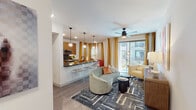 This thoughtfully designed single-floor residence combines modern aesthetics with functional living across a well-appointed open layout. The living and dining area is anchored by wide-plank wood-style flooring, a ceiling fan, and large sliding glass doors that invite abundant natural light and provide direct access to an outdoor balcony. Bold golden-yellow drapes frame the view, adding warmth and vibrancy to the space. A curved sage-green sofa and geometric area rug complete the contemporary living zone.

The kitchen opens seamlessly to the living area via a sleek breakfast bar with stylish striped stools. Warm wood cabinetry, dark countertops, a gray tile backsplash, and a full suite of stainless steel appliances — including a gas range, microwave, and refrigerator — make this kitchen as functional as it is attractive. Pendant globe lights add a refined finishing touch.

The bedroom is a bold, design-forward retreat featuring a richly upholstered yellow headboard, vibrant abstract wall art, ornate gold-framed mirrors, and dramatic patterned drapery. A ceiling fan and carpeted flooring contribute to a comfortable atmosphere.

The bathroom is clean and modern, featuring a tub-shower combination with white tile surround, wood-toned cabinetry, and dark countertops. A dedicated in-unit laundry area with a full-size washer and dryer rounds out this well-equipped residence.