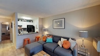 This single-floor residence offers a thoughtfully designed open-concept layout that seamlessly blends the kitchen and living areas into one cohesive, inviting space. The living room is anchored by a spacious sectional sofa accented with vibrant orange and teal throw pillows, adding a lively pop of color against the neutral gray walls. A textured ceiling and warm hardwood-style flooring contribute to the room's comfortable, lived-in atmosphere. Framed wall art and a softly glowing table lamp enhance the space with tasteful decorative touches. Adjacent to the living area, the kitchen features white cabinetry, dark countertops, and modern black appliances, including a full-size refrigerator. A breakfast bar with orange bar stools bridges the kitchen and living space, making it ideal for casual dining or entertaining. Under-cabinet lighting and a clean, minimal aesthetic give the kitchen a contemporary feel. The open flow between rooms maximizes the sense of space throughout the floor plan, making this residence feel bright, functional, and welcoming. Overall, the space strikes a balance between modern design and cozy livability.