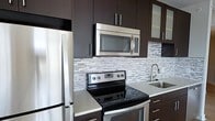 This two-floor space includes a thoughtfully designed kitchen that exemplifies modern style. The kitchen is outfitted with a full suite of stainless steel appliances, including a refrigerator, electric range with a smooth glass cooktop, over-the-range microwave, and a stainless steel sink. Dark espresso-toned cabinetry extends from the countertops to the ceiling, maximizing storage while lending a sleek, contemporary aesthetic. The countertops feature a light-toned surface that contrasts elegantly with the dark cabinetry. A striking mosaic tile backsplash in white, grey, and charcoal tones adds texture and visual interest throughout the cooking area. Frosted glass cabinet inserts near the upper cabinetry introduce a subtle decorative touch. The overall design of the kitchen emphasizes clean lines, functional layout, and a cohesive color palette that balances dark and light elements. Spanning two floors, the broader space offers versatile living arrangements, with the kitchen serving as a refined, well-equipped centerpiece of the home's interior design.