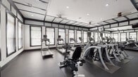 This dedicated fitness center is designed with both functionality and aesthetics in mind, presenting a sleek black-and-white color scheme that creates a sharp, professional atmosphere. The space is anchored by a robust selection of cardio equipment, including multiple treadmills, elliptical trainers, stair climbers, and upright and recumbent stationary bikes — many equipped with individual monitor screens for an enhanced workout experience. Large windows line the perimeter walls, fitted with blinds that allow ample natural light to fill the room while maintaining privacy. Ceiling fans complement the recessed lighting overhead, ensuring a comfortable environment during intense workouts. Full-length mirrors extend along one wall, visually expanding the space and allowing users to monitor their form. The flooring consists of durable dark rubber gym matting, providing both safety and sound absorption throughout. A storage area along one wall offers cubbies and shelving for personal belongings, while a weight and exercise accessory section — including stability balls and weight plates — supports a variety of training routines. Glass-paneled doors lead directly to an outdoor courtyard area with lush greenery, offering a refreshing connection to the outdoors.