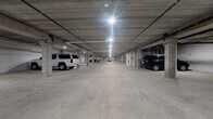 This single-level indoor parking garage is a well-structured, expansive facility designed to accommodate a large number of vehicles with ease and efficiency. The garage is built with a robust concrete construction throughout, featuring exposed concrete ceilings, structural support columns, and a smooth, light-colored concrete floor with clearly marked yellow striping to delineate individual parking stalls and traffic lanes.

The stalls are individually numbered along the perimeter walls, allowing for straightforward identification and organization. Dedicated ADA-accessible parking spaces are present, marked with the standard blue signage and floor markings, ensuring compliance and convenience for all users.

Overhead, a grid of bright LED high-bay light fixtures runs the length of the facility, providing consistent and ample illumination across the entire floor. Utility infrastructure, including piping and conduit runs, is visible along the ceiling, characteristic of a functional, no-frills commercial parking structure.

Wide central drive aisles offer easy maneuverability for vehicles of all sizes, including full-size trucks and SUVs. Exit signage and fire safety equipment, including fire extinguishers, are strategically positioned throughout. The overall ambiance is clean, orderly, and well-maintained, making this garage a practical and reliable parking solution.