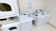 This single-floor communal laundry room offers a practical and well-organized space designed to meet shared laundry needs efficiently. The room is outfitted with multiple commercial-grade top-loading washing machines and a stacked dryer unit, all finished in crisp white and arranged to maximize accessibility and workflow. The machines appear to be coin- or card-operated, catering to a shared-use environment. A utility sink is conveniently positioned between the machines, providing an added resource for hand-washing or pre-treating garments. The walls are painted in a clean, neutral tone, contributing to the bright and tidy atmosphere of the room. Informational signage is posted on the wall, offering laundry service guidelines for users. The flooring is light-colored and easy to maintain, reinforcing the overall cleanliness of the space. The layout is straightforward and functional, allowing multiple users to operate machines simultaneously without congestion. This laundry facility is well-suited for a residential building or similar shared living environment, providing residents with reliable and accessible laundry amenities in a neatly kept setting.
