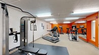 Located at 140 Lavale Dr in Monroeville, PA, this fitness facility offers a single-floor exercise room designed to support a well-rounded workout routine. The space is efficiently laid out to maximize usability, featuring a mix of cardio and strength training equipment that includes treadmills, stationary bikes, an elliptical, a stair climber, a multi-function cable machine, and a weight bench — catering to a variety of fitness goals.

The room is finished with durable, easy-to-clean flooring, complemented by interlocking rubber mat sections beneath the strength equipment for added safety and stability. A bold orange accent wall adds energy and visual interest to the space, while a large wall-mounted mirror spans one side of the room, enhancing the sense of openness and allowing users to monitor their form during workouts.

Overhead fluorescent lighting ensures the room is well-lit throughout. Instructional fitness posters are displayed on the walls, offering exercise guidance. A small seating area near the entrance provides a convenient spot to rest, and wall-mounted amenities such as a paper towel dispenser contribute to the room's practicality. The drop-panel ceiling and clean white walls give the space a tidy, functional appearance suited for regular fitness use.