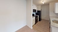This single-floor unit features a thoughtfully designed open-concept layout that seamlessly connects the living and kitchen areas. The living space is carpeted in neutral tones and benefits from natural light through a large window, complemented by a ceiling fan with integrated lighting. A peninsula with granite countertops and pendant lighting serves as a natural divider between the living area and the kitchen, which transitions to durable wood-look flooring.

The kitchen is well-appointed with white shaker-style cabinetry featuring brushed nickel hardware, granite countertops, a classic white subway tile backsplash, and black appliances including a refrigerator, range, microwave, and dishwasher. Upper open shelving adds additional storage and visual interest.

The bedroom is generously sized with plush carpeting, dual windows that invite natural light, and a ceiling fan overhead. The bathroom includes a full soaking tub with an integrated shower, white tiled surround with built-in corner shelving, and chrome fixtures. Throughout the unit, a consistent neutral color palette and clean finishes create a bright, cohesive, and functional living environment.