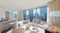 This sophisticated high-rise residence offers a thoughtfully designed open-concept floor plan bathed in abundant natural light. The living area is anchored by dramatic floor-to-ceiling curved windows that frame breathtaking panoramic city skyline views, creating a striking visual centerpiece. Light hardwood flooring flows throughout the main living spaces, complementing the neutral, modern palette.

The kitchen is outfitted with light wood cabinetry, quartz countertops, stainless steel appliances including a built-in oven and gas cooktop, and a generous central island with an integrated sink — seamlessly connecting to the dining and living areas.

A versatile den or study provides a quiet retreat, featuring light wood flooring, a dedicated workspace, and a comfortable seating area. The primary bedroom is a serene sanctuary with direct city views through floor-to-ceiling windows, with an en-suite bathroom showcasing light wood vanity cabinetry, quartz countertops, and a large illuminated mirror. A second full bathroom features a sleek, all-white double-vanity design. The second bedroom also enjoys elevated city views. Throughout, the residence maintains a cohesive, contemporary aesthetic defined by clean lines, light tones, and high-quality finishes.