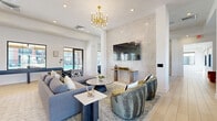 Located in Buckeye, Arizona, this thoughtfully designed single-floor amenity space offers a sophisticated blend of social, recreational, and professional environments. The open-concept main lounge is anchored by soaring ceilings, an eye-catching gold sputnik chandelier, and a dramatic floor-to-ceiling tiled accent wall that frames a large wall-mounted television. Plush sectional sofas, accent chairs, and layered area rugs create an inviting atmosphere throughout the lounge. Light wood-look flooring and hexagonal tile work together to define distinct zones within the space, while large black-framed windows and glass doors flood the interior with abundant natural light and provide views of the outdoor pool area. A wet bar with counter seating and woven pendant lighting serves as a social hub, ideal for gatherings. The game room features a full-size billiards table set beneath statement rattan pendants, complemented by bold artwork and a tiled accent wall. A wide hallway connects all areas and includes a shuffleboard table alongside direct access to the outdoor amenities. Two dedicated office spaces provide functional workstations, built-in cabinetry, wall-mounted displays, and pool-facing windows, offering a productive and well-lit environment for focused work.
