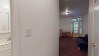 This single-floor unit offers a straightforward and functional layout designed to maximize the use of its available space. The main living and dining area is carpeted in a deep burgundy tone and is illuminated by a ceiling-mounted light fixture. A large window dressed with sheer curtains allows natural light to filter into the room, creating a bright and welcoming atmosphere. The space comfortably accommodates a small round wooden dining table with chairs as well as a dark upholstered futon, making it suitable for both relaxing and dining.

A carpeted hallway connects the main living area to the rest of the unit, leading to a closet and additional rooms. The kitchenette is efficiently arranged with warm-toned wooden cabinetry both above and below the counter, a white electric range, a single-basin sink, and a full-size white refrigerator. The flooring in the kitchen area transitions to a durable tile pattern, distinguishing it from the carpeted living space.

The bathroom is clean and simply appointed, featuring a single-sink vanity with under-cabinet storage, a tri-panel mirrored medicine cabinet above, and a full-size bathtub with a showerhead. The all-white color palette throughout the bathroom contributes to a bright, tidy feel. Overall, the unit presents a practical and cohesive living environment suitable for individuals or couples.