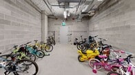 This dedicated bicycle storage room is located within the building at 2540–2590 Lonsdale Avenue in North Vancouver, BC. The space is constructed with exposed concrete block walls and a poured concrete floor, giving it a durable, utilitarian character well-suited to its purpose. The room spans a generous single level, offering enough open floor area to accommodate a large number of bicycles of varying sizes — from adult mountain bikes and commuter bikes to children's bikes and ride-on toys.

Overhead, the ceiling features exposed mechanical infrastructure including piping, conduit runs, and fluorescent strip lighting, which provides even illumination throughout the space. A fire alarm system is visibly installed, and a clearly marked emergency exit door is accessible at one end of the room. Ventilation units are mounted along the upper walls, ensuring adequate airflow within the enclosed space.

Bike racks are positioned throughout the room, allowing for organized storage along the perimeter and in the central area. The layout accommodates a high volume of bicycles while maintaining clear circulation paths. The secure, self-contained design of this storage room makes it a practical and functional amenity, particularly well-suited for residents in an urban area like North Vancouver where cycling is a popular mode of transportation.