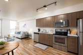 This modern single-floor unit is designed with a clean, contemporary aesthetic and an efficient open-concept layout. The living and kitchen areas flow seamlessly together, anchored by warm hardwood-style flooring that runs throughout the main space. The kitchen is outfitted with medium-toned wood cabinetry, stainless steel appliances including a refrigerator, electric range, microwave, and dishwasher, all complemented by light countertops and track lighting overhead. A breakfast bar area with stools provides an additional casual dining option.

The living room is bright and welcoming, benefiting from large windows and a glass-panel door that invite abundant natural light and offer views of the surrounding exterior. The space comfortably accommodates a seating arrangement centered around a glass coffee table, creating a relaxed yet functional gathering area.

The bedroom is a tranquil retreat featuring plush carpeting, a large window with outdoor views, and direct access to a generously sized walk-in closet with built-in shelving and hanging space. The bathroom features a wide vanity with ample counter space, wood-tone cabinetry, and a shower with curtain enclosure. A stacked washer and dryer unit is conveniently integrated into the bathroom, providing in-unit laundry accessibility. Overall, the unit offers a well-appointed, functional living environment with modern finishes throughout.