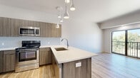 Located at 1677 Midtown Pl in Midwest City, Oklahoma, this modern single-floor residence showcases a clean, contemporary design throughout. Light wood-tone flooring runs consistently across the entire unit, creating a bright and cohesive aesthetic.

The open-concept kitchen is a standout feature, equipped with stainless steel appliances including a refrigerator, range, and over-the-range microwave. Warm wood-grain cabinetry pairs with a geometric hexagon tile backsplash and a large quartz-topped island with an undermount sink, offering both functionality and style. Pendant lighting hangs above the island, adding a modern finishing touch.

The living area flows naturally from the kitchen and opens to a private balcony through sliding glass doors, providing pleasant outdoor views of surrounding trees. Multiple additional rooms, suitable for bedrooms or flex spaces, feature large windows that invite generous natural light and frame lush green tree canopy views. One room includes a sleek barn-style sliding door for added character.

The bathroom features a glass-enclosed walk-in shower with textured white tile surround, a built-in niche, and brushed nickel hardware, reflecting the unit's modern sensibility. Throughout the space, clean white walls and understated finishes create a bright, move-in-ready environment.