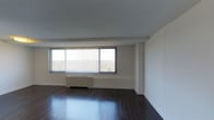 This single-floor space presents a clean, minimalist aesthetic that emphasizes openness and natural light. The main living area is anchored by rich, dark hardwood flooring that contrasts sharply with the crisp white walls and ceiling, creating a sleek, modern look. A bank of large windows dominates one wall, fitted with horizontal blinds that allow for adjustable light control. These expansive windows frame broad outdoor views, suggesting an elevated position that enhances the sense of openness throughout the room. A baseboard heating unit sits beneath the windows, integrated neatly into the space without disrupting the clean lines of the interior. The walls are smooth and neutral, offering a versatile backdrop suitable for a variety of furnishing styles. The ceiling is flat and uniformly white, contributing to the room's airy, uncluttered feel. The overall ambiance is calm and functional, with an emphasis on simplicity and practicality. The generous floor area provides ample room for flexible furniture arrangements, making this space well-suited for use as a comfortable living area. The combination of natural light, polished flooring, and open layout gives the space a welcoming and contemporary character.