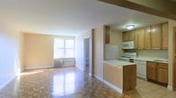 Located at 44-30 Douglaston Pkwy in the quiet, residential neighborhood of Douglaston, Queens, this single-floor unit offers a practical and welcoming layout. The open-plan living and kitchen area is the heart of the space, bathed in natural light from a large window that frames views of the surrounding greenery. The living area features classic parquet hardwood flooring with a polished finish that adds warmth and character to the room. A wall-mounted air conditioning unit ensures year-round comfort. The kitchen is open to the living space and designed for functionality, featuring warm wood cabinetry, a gas range, a microwave, a refrigerator, and ample counter space. A peninsula counter serves as a natural divider between the kitchen and living areas, offering additional workspace and casual seating potential. A structural beam detail at the ceiling adds architectural interest to the open layout. A doorway visible from the main living area leads to additional rooms, providing a sense of depth and connectivity throughout the unit. Clean white trim and neutral wall tones throughout create a bright, cohesive atmosphere that makes the space feel open and move-in ready.
