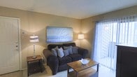This single-floor space offers a comfortable and well-appointed interior designed with a warm, neutral color palette that creates an inviting atmosphere throughout. The living area serves as the heart of the space, centered around a plush, dark upholstered sofa accented with decorative pillows, complemented by rich wooden side and coffee tables that add a grounded, earthy feel. Ambient lighting from stylish table and floor lamps enhances the cozy ambiance, casting a soft warm glow across the room. A large framed nature-inspired wall art piece depicting coastal grasses adds a serene focal point above the sofa, contributing to the calm and relaxed tone of the space. The textured ceiling adds subtle architectural character, while carpeted flooring underfoot provides comfort. A sliding glass door dressed with sheer curtains allows natural light to filter gently into the room, brightening the space while maintaining a sense of privacy. The overall design strikes a balance between functional living and understated elegance, making the space feel both practical and welcoming for everyday use.