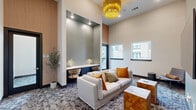 Located at 4114 FM 2920 Road in Spring, Texas, this thoughtfully designed lounge space combines modern aesthetics with functional comfort. The single-floor layout maximizes every square foot, anchored by a dramatic soaring ceiling height that lends an open, airy atmosphere well beyond its physical footprint.

The living area serves as the heart of the space, centered around a plush white sofa accented with warm earth-tone throw pillows. A wall-mounted flat-screen television faces the seating area, while rustic wood cube side tables and a sleek modern chaise longue round out the furnishings. Overhead, a striking gold chandelier with cascading fringe serves as a bold focal point, adding a touch of glamour to the refined interior.

A built-in workstation is tucked neatly into a recessed niche along one wall, featuring a marble-topped desk surface, navy-blue cabinetry, and elegant brass task lighting — ideal for focused work or study. A patterned geometric carpet in muted tones anchors the room and complements the neutral warm-grey and sage color palette throughout.

Natural light filters in through multiple windows, offering views of the surrounding exterior. Warm wood-panel wall accents and dark-framed glass doors add architectural depth and contrast, while recessed lighting provides a clean, polished finish. The overall ambiance is sophisticated yet inviting.