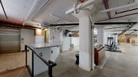 Located at 12 S 17th St in Richmond, VA, this expansive exercise room occupies a single floor and offers a generous, open layout designed to accommodate a wide range of fitness activities. The space showcases a compelling industrial-chic aesthetic, defined by exposed brick walls, polished concrete floors, and an open ceiling with visible ductwork, piping, and structural beams painted white — all contributing to a raw yet refined ambiance.

The fitness area is comprehensively equipped, featuring strength training machines, cable systems, rowing machines, ellipticals, stationary bikes, free weights, medicine balls, TRX suspension trainers, and stretching areas with floor mats. Structural columns punctuate the floor plan, adding architectural character while naturally delineating zones within the space. Locker banks provide convenient storage for users.

A kitchenette with a granite countertop and utility sink serves as a functional support area, complemented by a casual lounge with a leather sofa and area rug. Additional rooms accessible via clean white-paneled doors offer further utility. Small windows along the brick walls allow modest natural light to filter in. The overall atmosphere is purposeful and energetic, blending historic building character with modern fitness functionality.