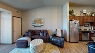 This single-floor residence offers a thoughtfully designed layout that balances comfort and functionality. The open living area features light wood-style flooring, vaulted ceiling accents, and a sliding glass door that floods the space with natural light while providing views of the surrounding greenery outdoors. The living room comfortably accommodates seating and flows seamlessly into the kitchen area.

The kitchen is equipped with rich wood cabinetry, dark countertops, and a full suite of stainless steel appliances including a refrigerator, dishwasher, range with over-the-range microwave, and ample storage space. A dedicated mudroom entry near the kitchen offers a practical transition space from the exterior door.

The bedroom is a standout feature of the home, showcasing plush carpeting, a sliding barn-style closet door, and a striking crystal chandelier that adds an elegant, decorative touch. A window dressed with layered curtains allows soft natural light to filter in, creating a warm and restful ambiance.

The full bathroom is outfitted with a wood-tone vanity, a wide mirror with overhead lighting, and a shower-tub combination. Light wood-style flooring continues into this space, maintaining a cohesive aesthetic throughout the home. Overall, the residence presents a welcoming and well-maintained interior with consistent warm-toned finishes.