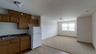 This single-floor space showcases an open layout that seamlessly connects the kitchen and living area, creating a functional and cohesive environment. The kitchen is equipped with warm medium-toned wood cabinetry, offering generous upper and lower storage. A white refrigerator anchors the kitchen zone, complemented by a dark countertop that provides a subtle contrast to the lighter surroundings. The flooring transitions from smooth light-toned hardwood in the kitchen area to plush neutral carpeting in the living space, clearly defining each zone while maintaining a unified aesthetic. The living area benefits from a window that allows natural light to fill the room, brightening the white walls and contributing to an airy, open atmosphere. Ceiling-mounted light fixtures provide additional illumination throughout the space. The clean, neutral color palette of white walls paired with the wood-toned cabinetry and mixed flooring creates a practical yet inviting interior that is well-suited for everyday living.