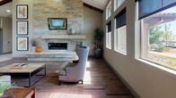 This single-floor community space is designed with an open, airy layout that immediately conveys a sense of scale and sophistication. The entrance features large glass doors framed in warm taupe finishes, with transom windows above that draw in abundant natural light and offer views through to the interior and surrounding outdoor areas. Exposed dark wood beams traverse the high ceilings throughout, adding rustic character to the otherwise contemporary design.

The main lounge area is anchored by a striking stacked-stone fireplace wall with a built-in television above a clean, floating mantel. Elegant high-back wing chairs and area rugs are arranged thoughtfully on rich wood-look flooring, creating an inviting atmosphere. Tall banks of windows along one wall flood the space with daylight while framing outdoor views of the surrounding community.

A dedicated kitchenette area features dark walnut cabinetry with dark granite countertops, stainless steel appliances including a full-size refrigerator and warming drawer, and decorative wall-mounted accents that lend a distinctive, lodge-inspired character to the space.

Large glass doors on the opposite side of the main hall open directly to an outdoor patio, seamlessly connecting interior and exterior living areas. Light tile flooring in the entry and transitional corridors contrasts with the warm wood tones of the lounge, creating a well-defined and cohesive flow throughout.