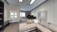 This single-story clubhouse is designed to serve as a functional and comfortable community gathering space. The interior features a neutral color palette of grey and taupe tones, complemented by patterned carpeting throughout and vaulted ceiling elements that lend a sense of openness and height to the main area.

The lounge zone is furnished with a large sectional sofa and accent chairs arranged around a substantial dark bookshelf unit housing a television, creating a comfortable entertainment area. Decorative wall art in yellow and grey tones adds visual interest throughout the space. A water cooler is conveniently positioned nearby.

A dedicated workstation area includes a dark wood desk, computer equipment, and an ergonomic office chair, providing residents with a functional space for business or personal tasks. Natural light filters in through multiple windows fitted with blinds, keeping the space bright and welcoming.

A wooden dining or activity table is also present, adding to the multi-use nature of the clubhouse. Additional rooms are accessible through interior doors, suggesting supplementary spaces for varied community needs. Recessed and skylight-style ceiling fixtures provide ample artificial lighting throughout.