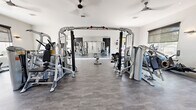 This fitness center is a single-floor facility designed to accommodate a full spectrum of workout needs in a clean, modern environment. The space is anchored by a spacious open floor plan finished with durable grey wood-look flooring that provides both functionality and a contemporary aesthetic. Crisp white walls and recessed lighting are complemented by multiple ceiling fans, ensuring the space remains well-ventilated and comfortable during exercise.

At the heart of the facility is a multi-station cable machine system, surrounded by a variety of selectorized strength machines from Hoist, catering to a wide range of muscle groups. Adjustable weight benches are positioned throughout for free-weight and functional training exercises. A dedicated cardio zone features multiple treadmills positioned beside large windows with roller shades, allowing natural light and outdoor views while working out.

Full-length wall mirrors are strategically installed to maximize the sense of space and allow users to monitor their form. An accent wall with a bold, graphic patterned wallcovering adds a modern design touch, flanked by open wooden shelving for storing accessories such as exercise balls. A glass door provides direct outdoor access, adding to the facility's bright, airy ambiance. The overall layout is open, well-organized, and thoughtfully designed for an efficient and motivating fitness experience.