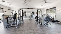 This fitness center is a single-floor facility designed to accommodate a full spectrum of workout needs in a clean, modern environment. The space is anchored by a spacious open floor plan finished with durable grey wood-look flooring that provides both functionality and a contemporary aesthetic. Crisp white walls and recessed lighting are complemented by multiple ceiling fans, ensuring the space remains well-ventilated and comfortable during exercise.

At the heart of the facility is a multi-station cable machine system, surrounded by a variety of selectorized strength machines from Hoist, catering to a wide range of muscle groups. Adjustable weight benches are positioned throughout for free-weight and functional training exercises. A dedicated cardio zone features multiple treadmills positioned beside large windows with roller shades, allowing natural light and outdoor views while working out.

Full-length wall mirrors are strategically installed to maximize the sense of space and allow users to monitor their form. An accent wall with a bold, graphic patterned wallcovering adds a modern design touch, flanked by open wooden shelving for storing accessories such as exercise balls. A glass door provides direct outdoor access, adding to the facility's bright, airy ambiance. The overall layout is open, well-organized, and thoughtfully designed for an efficient and motivating fitness experience.