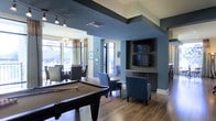This well-appointed single-floor amenity space combines entertainment and relaxation in a cohesive, modern design. The room is anchored by a full-size billiards table set atop rich hardwood-style flooring, making it an immediate focal point for social gatherings. Adjacent to the game area, a built-in media nook features dual wall-mounted flat-screen televisions framed within a deep charcoal accent wall, accompanied by comfortable blue upholstered lounge chairs that invite residents to unwind.

The space flows naturally into a dedicated dining and lounge area, distinguished by a tray ceiling detail and a decorative chandelier that adds a refined touch. Large floor-to-ceiling windows and glass doors line the perimeter, flooding the interior with abundant natural light while offering views of the surrounding outdoor greenery. Curtain panels in warm gold tones complement the cool blue and slate-gray wall palette, creating a balanced and inviting atmosphere.

Architectural details such as the bold dark ceiling beam, textured accent walls, and recessed lighting contribute to the space's contemporary character. The open layout encourages a seamless transition between gaming, lounging, and dining, making this clubroom a versatile and well-designed communal hub.