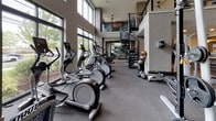 This expansive fitness center is thoughtfully designed to support a wide range of workout routines. The main floor is organized into distinct training zones, beginning with a generous cardio area lined with treadmills, elliptical machines, and stationary bikes arranged along floor-to-ceiling windows that flood the space with natural light and offer views of the surrounding greenery. Adjacent to the cardio section, a strength training area features cable machines, free weights, and resistance equipment, complemented by wall-to-wall mirrors that enhance the sense of depth throughout the space.

A functional training zone accommodates stability balls, resistance accessories, and open floor space for dynamic exercises. The lobby area features a sleek three-seat upholstered bench positioned beneath a large interior window overlooking the gym floor, alongside a textured stone accent wall with integrated media display—adding a refined, modern aesthetic to the entrance.

Double-height ceilings in key areas contribute to an airy, open atmosphere, further elevated by ceiling fans and recessed lighting. A striking spiral staircase connects the main floor to an upper mezzanine level, offering additional workout space. The neutral color palette, carpeted flooring, and modern architectural details create a polished, professional environment throughout.