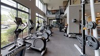 This expansive fitness center is thoughtfully designed to support a wide range of workout routines. The main floor is organized into distinct training zones, beginning with a generous cardio area lined with treadmills, elliptical machines, and stationary bikes arranged along floor-to-ceiling windows that flood the space with natural light and offer views of the surrounding greenery. Adjacent to the cardio section, a strength training area features cable machines, free weights, and resistance equipment, complemented by wall-to-wall mirrors that enhance the sense of depth throughout the space.

A functional training zone accommodates stability balls, resistance accessories, and open floor space for dynamic exercises. The lobby area features a sleek three-seat upholstered bench positioned beneath a large interior window overlooking the gym floor, alongside a textured stone accent wall with integrated media display—adding a refined, modern aesthetic to the entrance.

Double-height ceilings in key areas contribute to an airy, open atmosphere, further elevated by ceiling fans and recessed lighting. A striking spiral staircase connects the main floor to an upper mezzanine level, offering additional workout space. The neutral color palette, carpeted flooring, and modern architectural details create a polished, professional environment throughout.