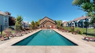 This outdoor amenity space centers around a large, rectangular lap pool with clear turquoise water, designed to accommodate both recreational swimming and exercise. The pool's elongated shape and smooth tiled surround give the area a clean, well-maintained aesthetic. Encircling the pool is a spacious concrete and paver sundeck, furnished with multiple wooden lounge chairs and benches positioned to take full advantage of open sky and abundant natural sunlight throughout the day.

A single-story pool house anchors one end of the space, featuring a distinctive peaked roofline with dark wood siding and red accent doors. The structure provides practical support facilities for pool users. The entire area is enclosed by a black wrought iron perimeter fence, ensuring both safety and a defined boundary within the broader residential community.

Mature trees and landscaped greenery line the outer edges of the fence, offering partial shade and a natural backdrop. Neighboring multi-story residential buildings with pitched roofs are visible beyond the fence line, contributing to a neighborhood atmosphere. The overall setting is bright, open, and well-suited for outdoor relaxation in warm weather.