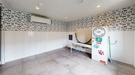 Located at 120 Commerce Way in Woburn, MA, this dedicated pet salon at The Point at Woburn is a purpose-built grooming space designed with both functionality and charm in mind. The room is finished with large-format neutral floor tiles that are easy to clean and maintain, while the lower half of the walls features classic white subway tile, providing a hygienic, water-resistant surface ideal for pet washing. Above the tile line, a whimsical wallpaper pattern featuring repeating dog silhouettes adds a playful, pet-friendly character to the space. The centerpiece of the room is a self-service dog wash station — a raised, walk-in style tub equipped with a hose and grooming attachments, making bath time convenient for pets of all sizes. A floor drain is centrally located to manage water efficiently. Recessed ceiling lighting provides bright, even illumination throughout, while a wall-mounted mini-split unit ensures year-round climate comfort. Plumbing connections are accessible along the tiled walls, and informational signage with pet salon rules is posted for user guidance. The space is accessed through a set of double exterior doors that allow ample natural light to enter, and a secondary interior door connects to an adjacent area. The overall design is clean, practical, and thoughtfully tailored to pet care.