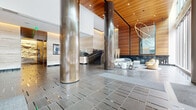 Located at 33 8th Street in San Francisco's SoMa district, this striking commercial lobby delivers a polished first impression through a refined blend of materials and thoughtful design. The expansive entry floor is laid in large-format stone tiles with metallic inlay detailing, reflecting the abundant natural light that pours through floor-to-ceiling glass facades facing the street. Towering cylindrical columns clad in brushed bronze anchor the open floor plan, drawing the eye upward toward the soaring double-height ceiling, which transitions from crisp white to warm, richly grained wood paneling — a defining architectural feature of the space. A marble-clad reception desk serves as the central point of arrival, set against a backdrop of travertine-style stone walls accented by integrated cove lighting. The lobby offers two distinct seating areas: one featuring a sculptural curved velvet sofa in soft grey paired with a marble accent table, and another with rounded, cognac leather lounge chairs arranged beside floor-to-ceiling windows overlooking the streetscape. A dramatic orbital chandelier adds an artistic focal point. Elevator banks with bronze-toned doors are set within travertine surrounds, while an open staircase with glass railings and brushed metal detailing connects to adjacent levels, completing this sophisticated urban lobby.