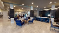 This single-floor amenity space is designed with a contemporary aesthetic that blends functionality with upscale comfort. The expansive main lounge serves as the heart of the space, featuring light travertine-style tile flooring, recessed lighting, and a layered tray ceiling with subtle accent lighting. Bold navy blue lounge chairs arranged around circular wood coffee tables create welcoming conversation areas, anchored by decorative area rugs. Striking dark wood-clad columns add architectural definition throughout the open floor plan.

A prominent front desk and bar-height counter with dark paneling provides a central service point, while nearby bench seating offers additional gathering space. The game room area features a pool table, arcade machines, and a shuffleboard table, all illuminated by elegant globe pendant chandeliers. Large windows allow natural light to fill the space.

A dedicated coffee bar nook showcases a professional-grade coffee machine, a utility sink, under-counter refrigeration, and textured tile wall accents, creating a cozy refreshment station. A self-serve market with stocked shelving is accessible nearby. The mail center features a well-lit room with extensive mailbox arrays, pendant lighting with gold interiors, and a central sorting counter, completing the range of resident-focused amenities in this thoughtfully designed space.