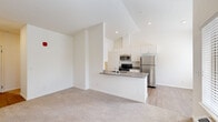 This single-floor residence offers a well-organized layout with distinct living, kitchen, and bedroom areas finished in a clean, neutral palette throughout. The open-concept living area features plush wall-to-wall carpeting and vaulted ceilings that lend a sense of spaciousness, with a large window overlooking greenery and an integrated climate control unit beneath it.

The kitchen is efficiently designed with white cabinetry extending to the ceiling for ample storage, dark countertops, and a full suite of stainless steel appliances including a refrigerator, range, microwave, and dishwasher. A peninsula counter separates the kitchen from the living area, providing both additional workspace and a natural visual boundary. Light wood-style flooring runs throughout the kitchen zone, contrasting with the carpeted living spaces.

The bedroom area features generous proportions, with vaulted ceilings contributing to an airy feel and a large window allowing natural light and outdoor views. A sliding closet offers organized storage with a built-in rod and shelf system. Additional rooms, visible in the 3D scan, include a secondary bedroom or flexible-use space with its own window and recessed lighting. Throughout the unit, white panel doors, recessed lighting, and a consistent neutral color scheme create a cohesive and functional living environment.