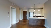 This contemporary single-floor residence showcases a thoughtfully designed open-concept layout that seamlessly blends living and kitchen spaces. Rich, dark hardwood floors run throughout, grounding the space with warmth and elegance. The kitchen is a standout feature, anchored by a substantial island with a textured concrete-style base and a clean white countertop, complemented by a stainless steel sink. White upper cabinetry with dark lower cabinets creates a striking two-tone contrast, while a white subway tile backsplash adds a classic touch. A stainless steel refrigerator and recessed lighting complete the functional kitchen setup. Three decorative pendant lights hang above the island, providing focused task lighting and a stylish focal point. The neutral wall tones in soft gray and warm beige promote a calm, versatile ambiance throughout. A glass-paneled door on one side adds a modern architectural detail, allowing light to flow between spaces, while a solid wood entry door with contemporary hardware anchors the entryway. The overall aesthetic is clean, modern, and move-in ready, combining durable materials with refined design choices for a polished everyday living experience.